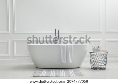 Modern ceramic bathtub and table with toiletries near white wall indoors Royalty-Free Stock Photo #2094777058