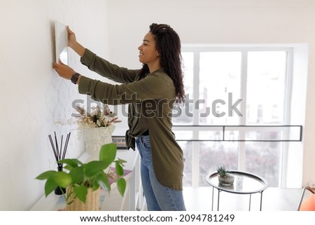 Modern Home Interior And Domestic Decor. Smiling young black woman hanging painting, putting photo picture frame on the wall. Casual lady taking care of coziness in her new stylish apartment, profile