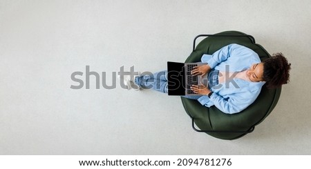People And Modern Technology. Above top view of young smiling black woman holding pc on lap sitting on bean bag in living room, typing on keyboard. Cheerful lady browsing internet, free copy space Royalty-Free Stock Photo #2094781276