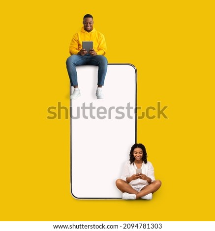 Great App. Excited Black Couple Sitting On Big Smartphone With Blank White Screen Using Gadgets, Cheerful Guy And Lady Chatting On Social Media, Yellow Orange Wall, Mock Up. Modern Communication Royalty-Free Stock Photo #2094781303
