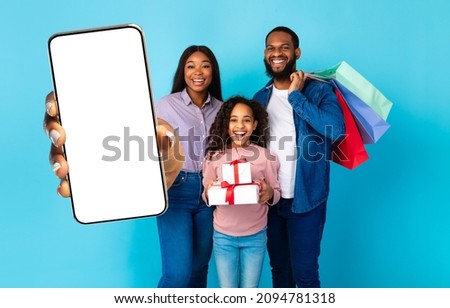 Mobile Shopping Application, Online Store. Happy Black Family Showing Blank Empty Cell Phone Screen Holding Shopper Bags And Gift Boxes Standing Isolated Over Blue Studio Background. Mock Up Template
