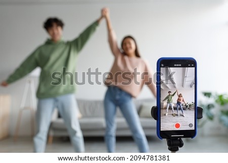 Couple of cheery Asian influencers shooting dance video for social network on cellphone, having fun at home, selective focus. Millennial bloggers streaming content for their vlog, broadcasting online Royalty-Free Stock Photo #2094781381