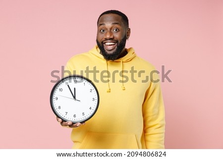 Excited cheerful young african american man 20s wearing casual yellow streetwear hoodie standing holding in hand clock looking camera isolated on pastel pink color wall background studio portrait