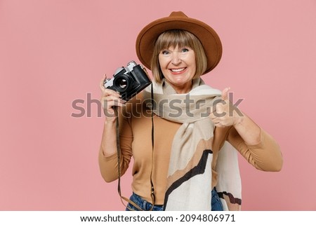 Traveler tourist mature elderly woman 55 years old wears brown shirt hat scarf hold retro vintage photo camera show thumb up like gesture isolated on plain pastel light pink background studio portrait