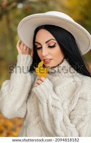 Fashion beautiful young girl with beauty face in beige vintage knitted sweater and hat holds a autumn yellow leaf and walks in the park. Female fall fashionable outfit look