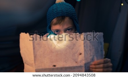 Close Up Shot of a Caucasian Young Boy Holding the Treasure Map and a Flashlight, Looking Serious As He Tries to Decipher the Code, Sitting and Hiding in a Dark Tent During The Night. Royalty-Free Stock Photo #2094843508