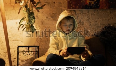 Cute Isolated Caucasian School Boy, Sitting on the Sofa of a Living Room, Holding a Smartphone, Watching a Movie on the Device. Wearing a Cozy White Hoodie, Background is An Industrial Apartment.
