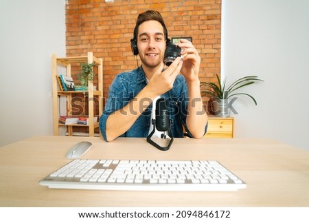 Front view of cheerful male blogger streaming live podcast recording review of camera lens using professional microphone sitting at desk, broadcasting at home office studio, looking at camera.