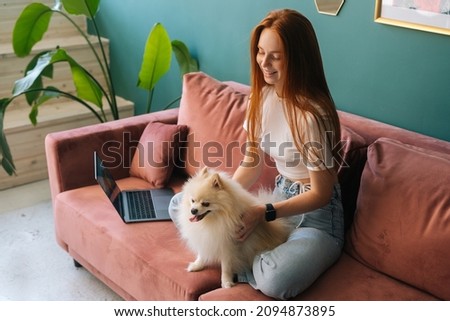 Side view of happy smiling young woman sitting on comfortable sofa cuddling pretty small white spitz pet dog at apartment. Cheerful female spending time with doggy at home during rest from laptop.