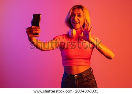 Greetings. Beautiful girl with headphones having videocall on her smartphone isolated over gradient background in neon lights. Concept of human emotions, facial expression, beauty. Copy space for ad