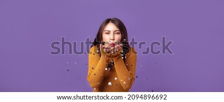 Young Asian woman cheerful blowing confetti on banner isolated purple studio background. Asian female happy in anniversary concept.
