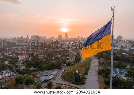 Aerial view of the Ukrainian flag waving in the wind against the city of Kyiv, Ukraine near the famous statue of Motherland at sunset. Royalty-Free Stock Photo #2094921034