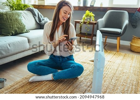 Woman dressed in beige sweater regulating heating temperature with a modern wireless thermostat and smart phone at home. Synchronization of thermostat with mobile devices concept