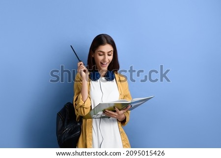 Happy broadly smiling student girl solved math problem, found clue, is looking at notebook and raising hand with pencil, wearing yellow shirt, white t-shirt, black bag and headphones over neck Royalty-Free Stock Photo #2095015426