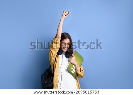 Happy student girl says yes, passed an important test, raises hand with fist in excitement, smiles sincerely, holding books, wearing yellow shirt, white t-shirt, black bag and headphones over neck