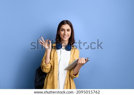 Happy smiling student girl is showing okay gesture with one hand and holding tablet with the other one, recommends application, wearing yellow shirt, white t-shirt, black bag and headphones over neck