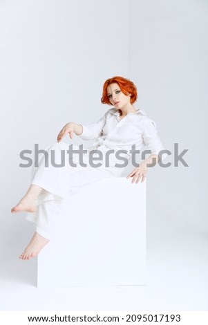beautiful young girl in the white t-shirt and pants with foxy hair posing on white cube on the white wall 