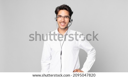 young hispanic man smiling happily with a hand on hip and confident. telemarketer concept Royalty-Free Stock Photo #2095047820