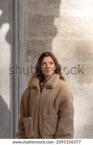 A young girl in front of the brown wooden door. She is looking up and far. She has warm clothes. Sunlight. Sunstroke to her face.