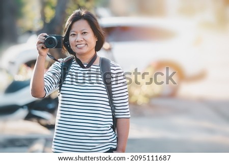 Happy Asian woman traveling and taking pictures outdoors city streets,
Portrait Asian female happy smiling backpacker using camera to take image on city walk streets, business travel concept.