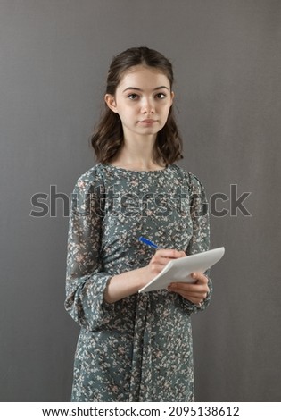 A beautiful successful girl in a dress stands against a gray wall with a notebook and pen in her hands