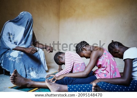 Totally covered person holding a blade speaking to a group of intimidated black African girls sitting in a row on the floor with their heads bowed prior to the female genital mutilation intervention Royalty-Free Stock Photo #2095148470