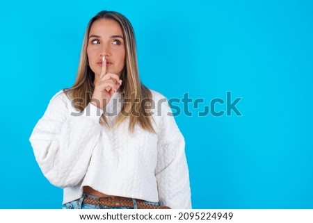 Young hispanic girl wearing white knitted sweater over blue background silence gesture keeps index finger to lips makes hush sign. Asks not to share secret