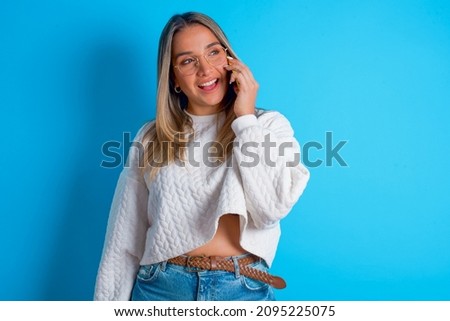 Pleasant looking happy Young hispanic girl wearing white knitted sweater over blue background has nice telephone conversation and looks aside, has nice mood and smiles positively while talks via cell 