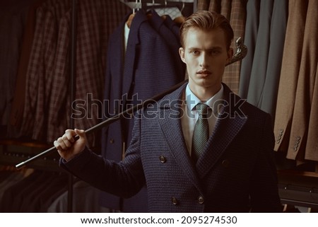 Fashion shot of a serious handsome blond man in elegant coat and three-piece suit with a walking stick in his hand standing in the background of luxury classic suits in a store. Men's fashion. Royalty-Free Stock Photo #2095274530