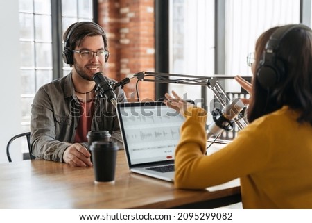 Smiling handsome man talking with gesturing pretty woman radio host while they recording live audio podcast in studio. Two people using laptop, headphones and audio equipment for making podcast