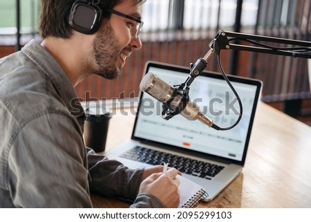 Close-up of man podcaster influencer blogger smiling while broadcasting his live audio podcast in studio using headphones, laptop and headphones. Male radio host in glasses making podcast or interview