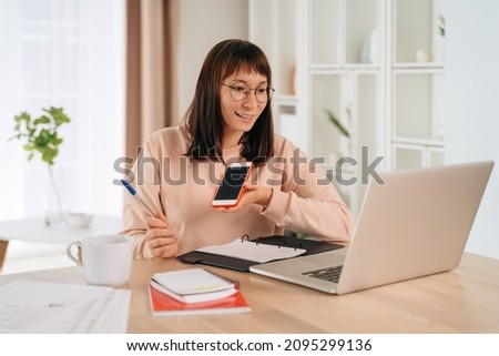 Beautiful young woman work remotely from home office, recording voice message on smartphone, using laptop, making notes, retraining. E-learning, distance education, re-skill and upskill concept Royalty-Free Stock Photo #2095299136