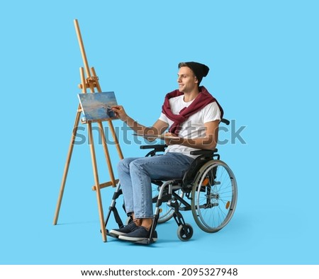 Young male artist in wheelchair painting picture against color background