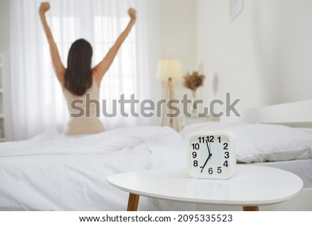 Good morning. Wake up. Rise and shine. It's time to start new working day. White alarm clock that tells 7 AM standing on bedside table, and woman who woke up from night sleep stretching in background Royalty-Free Stock Photo #2095335523