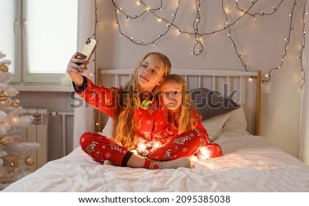 beautiful little girls are sitting in bed in bright Christmas pajamas taking pictures of themselves on the phone, a bright room.