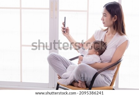 Beautiful Asian mother taking selfie picture by smartphone with daughter on chair. Women and baby sitting together and pay attention with camera. Todlder sitting on mother lap.