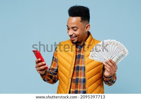 Excited fun young black man 20s years old wears yellow waistcoat shirt using mobile cell phone hold fan of cash money in dollar banknotes isolated on plain pastel light blue background studio portrait