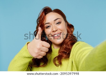 Young close up chubby overweight plus size big fat fit woman wear green sweater do selfie shot pov mobile phone show thumb up isolated on plain blue background studio portrait People lifestyle concept