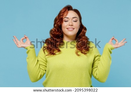 Young relaxed chubby overweight plus size big fat fit woman wear green sweater hold hands in yoga om aum gesture relax meditate calm down isolated on plain blue background. People lifestyle concept