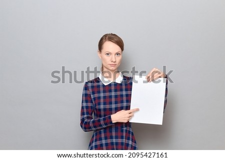 Studio portrait of serious young woman holding white blank paper sheet, pointing with index finger at empty place for your text and design, wearing checkered dress, standing over gray background