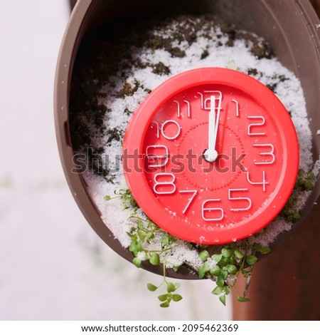 Red clock with white hands at 12 o'clock and 1 second with snowflakes on glass in a snow-covered flower pot with a green sprig of grass among the snow and black earth on a winter morning