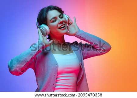 Happy young pretty girl in warm gray cardigan listening to music in headphones isolated on gradient blue orange neon background. Concept of emotions, facial expression, youth, studying, aspiration Royalty-Free Stock Photo #2095470358