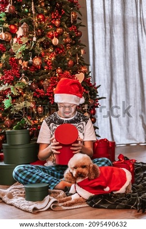 Cute teenage boy in Santa Claus hat is sitting under Christmas Tree with fluffy dog poodle pet on Christmas morning with gifts open wrapped presents, New Year celebration, family at home 