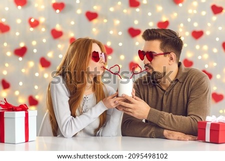 Young man and woman having a fun date on Saint Valentine's Day, sitting at table in cafe or at home, sipping drink from one cup through heart-shaped straws and enjoying cute and funny couple moment Royalty-Free Stock Photo #2095498102