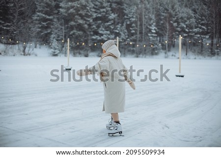 
A young girl in a white down jacket and a white hat is skating. Skating rink on the river against the background of a snowy winter forest