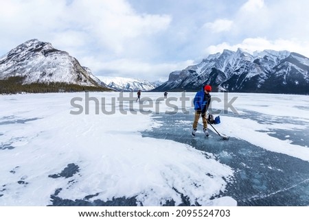 Boy practicing ice hockey on frozen Lake Minnewanka in the Canadian Rockies of Banff National Park during cold the Winter with unidentifiable visitors walking on the lake in the background.
