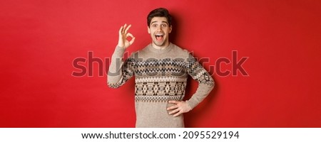 Concept of christmas celebration, winter holidays and lifestyle. Amazed and happy man in xmas sweater, showing okay sign and looking at something fantastic, red background