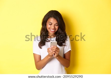 Portrait of good-looking african american girl in white t-shirt, messaging with smartphone, using mobile phone app, standing over yellow background