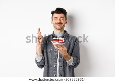 Hopeful birthday guy making wish with fingers crossed, holding bday cake on party, standing against white background