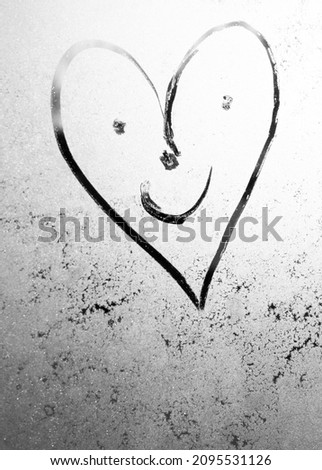heart with a drawn face with voices and a smile on frosty glass in winter with a finger, heart emoticon drawing by February 14 on misted glass, moonlight, texture of frost and water drops. out focus